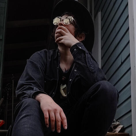 Fenrir Cerebellion dressed in black looking up at an overcast sky through bare tree branches reflected on their sunglasses.